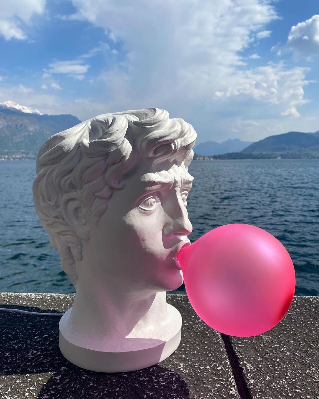 Outdoors on a stone ledge by a lake and mountains, this Clérachic bust lamp blows a bright pink bubble under a blue sky.