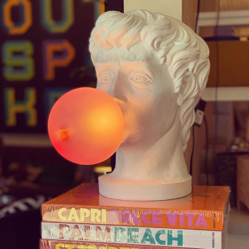 Close view of a Clérachic bust lamp with its bubble lit orange, standing on a pile of travel books against a bright backdrop.