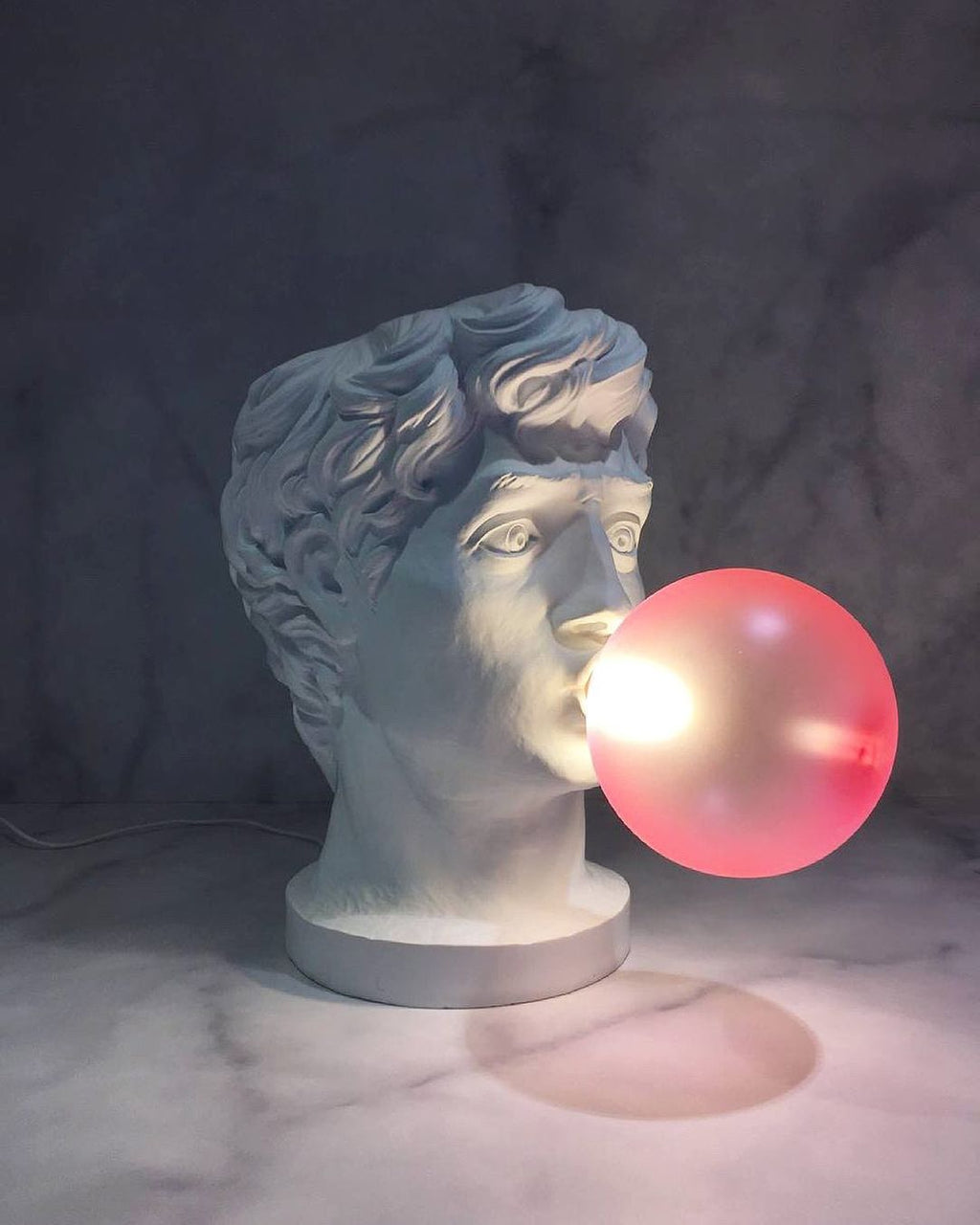A Clérachic bust lamp on a marble surface with a softly lit pink bubble, the face cool grey against a dark wall.