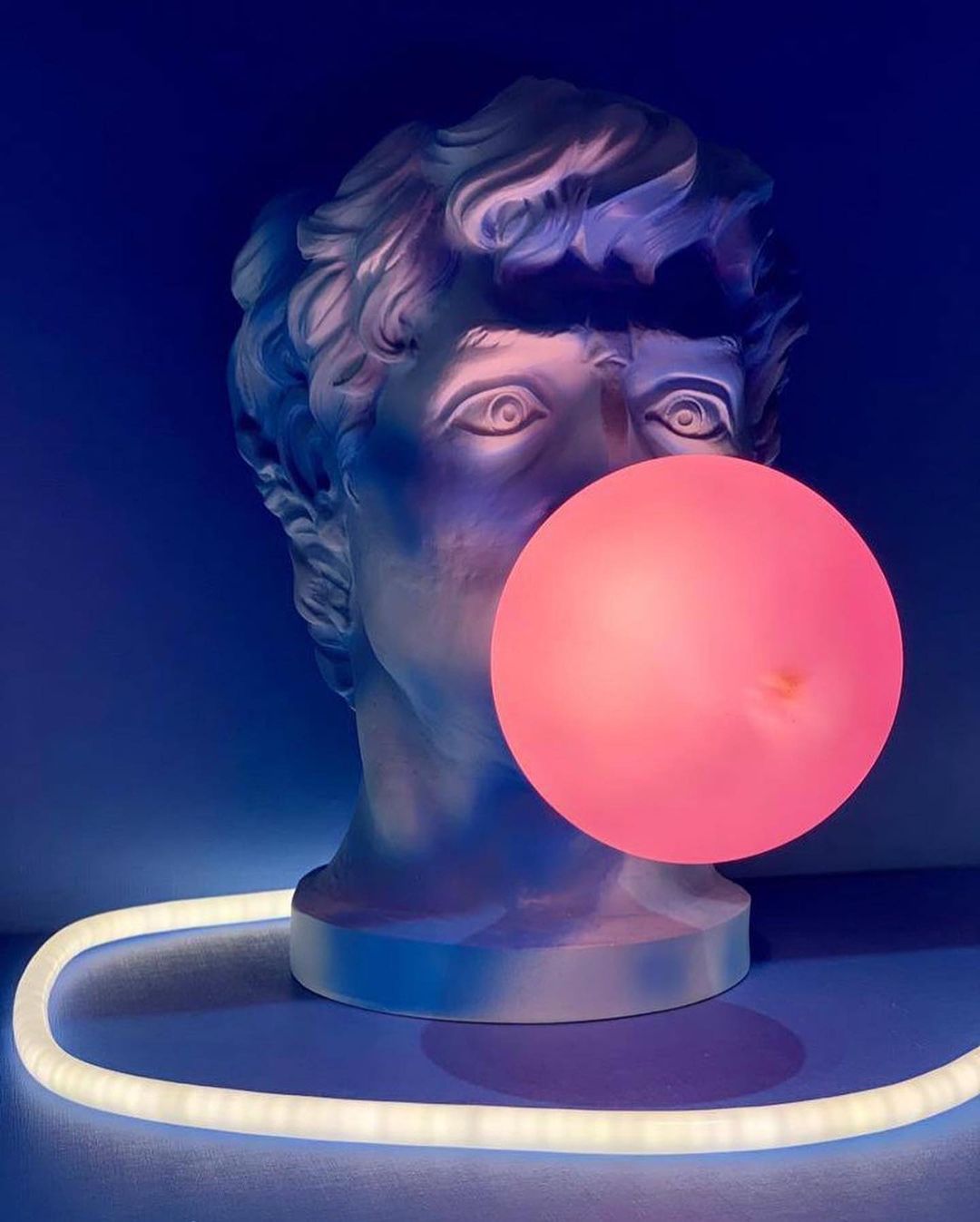 A Clérachic bust lamp glows in blue light with a pink-red bubble, and a ring of white light on the floor around its base.