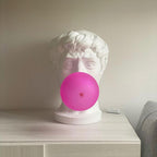 Seen from the front, this Clérachic bust lamp has a bold pink bubble covering the mouth, standing on a pale chest of drawers.