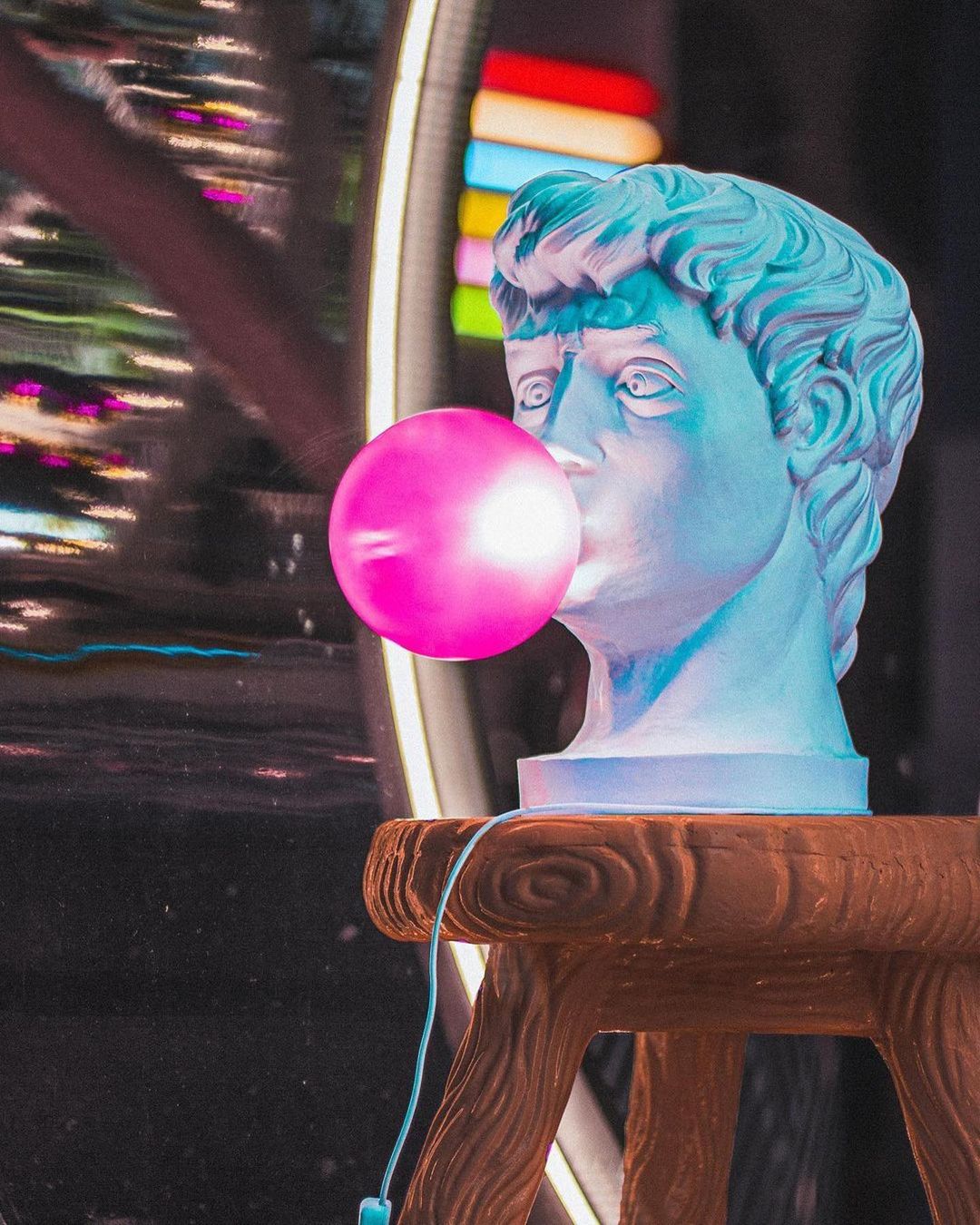 A Clérachic bust lamp with a lit pink bubble stands on a carved wooden stool in a dark room with neon signs in the background.