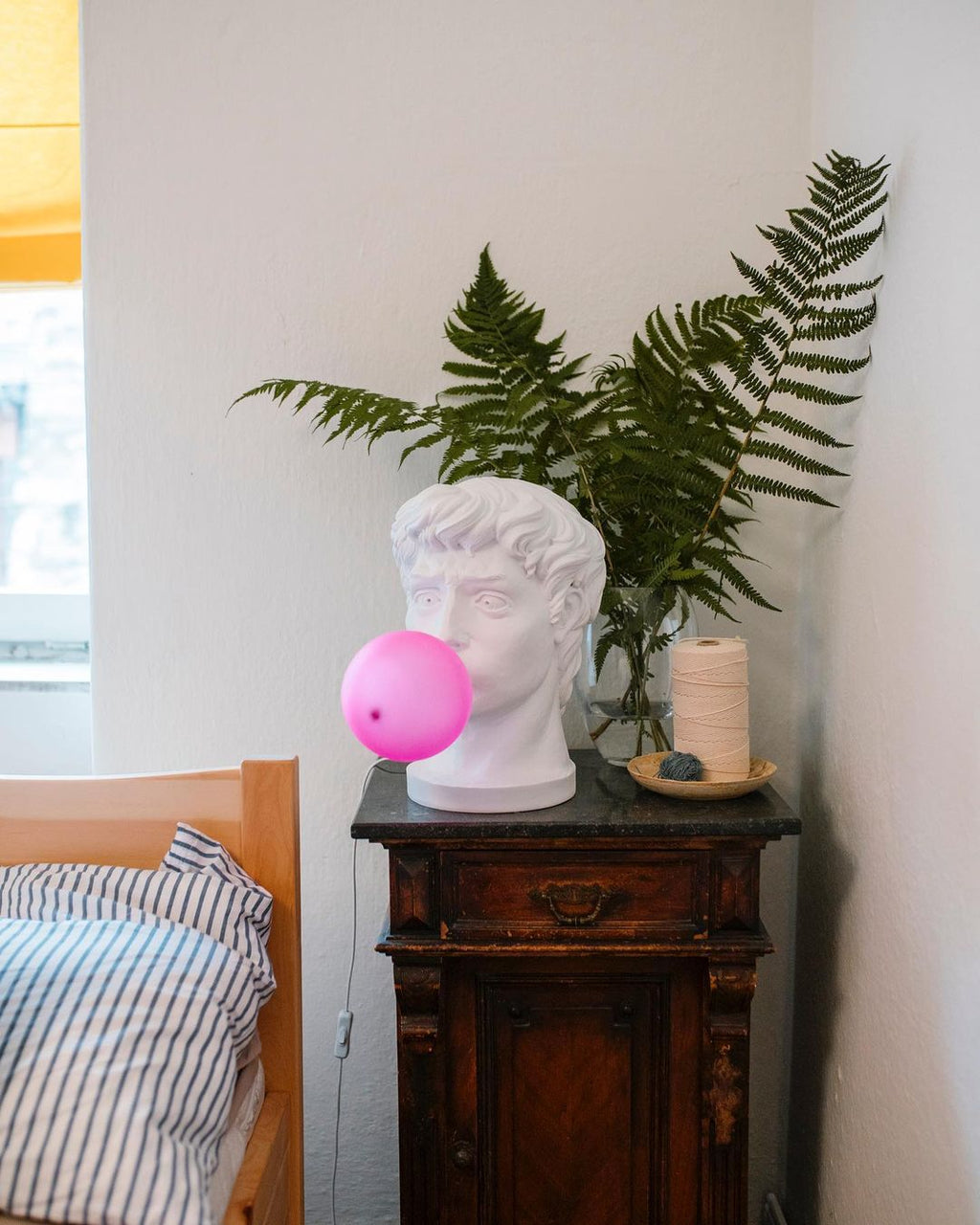 A Clérachic bust lamp with a small pink bubble stands on a dark cabinet next to a fern, with a bed beside it.