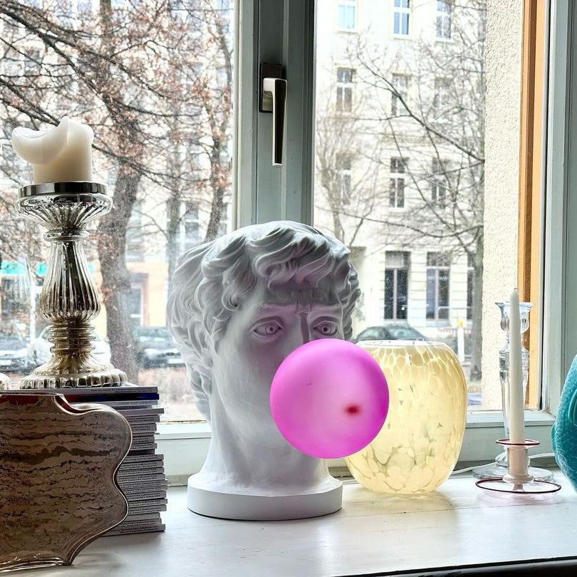 On a windowsill with candlesticks and a glass vase, this Clérachic bust lamp has a bright magenta bubble, with bare trees outside.