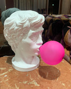 A Clérachic bust lamp with a bold pink bubble stands on a marbled wooden table, its shadow falling to the right.