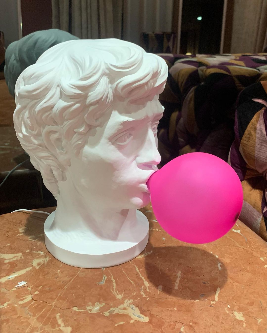 A Clérachic bust lamp with a bold pink bubble stands on a marbled wooden table, its shadow falling to the right.