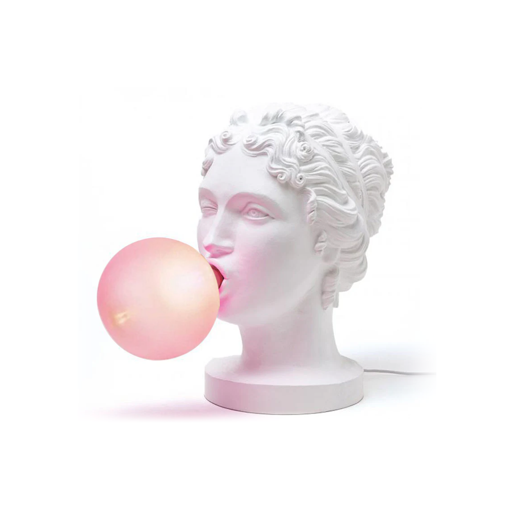 A Clérachic female bust lamp in white, blowing a pale pink bubble, on a plain white backdrop with a thin white cord.