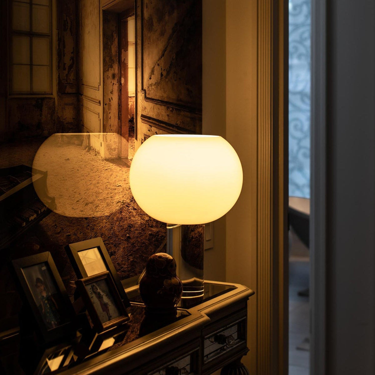 Glowing white glass globe of this Clérachic table lamp on an ornate console, with framed photographs and a moody painted room behind.