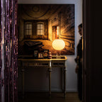 A lit Clérachic table lamp on a gilt-edged console table beneath a large painting, with a purple beaded curtain at the left.