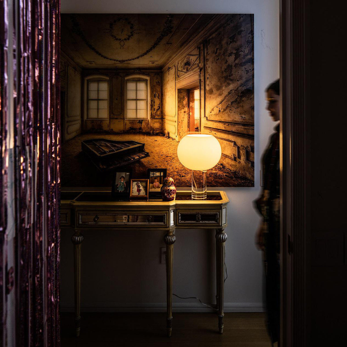 A lit Clérachic table lamp on a gilt-edged console table beneath a large painting, with a purple beaded curtain at the left.