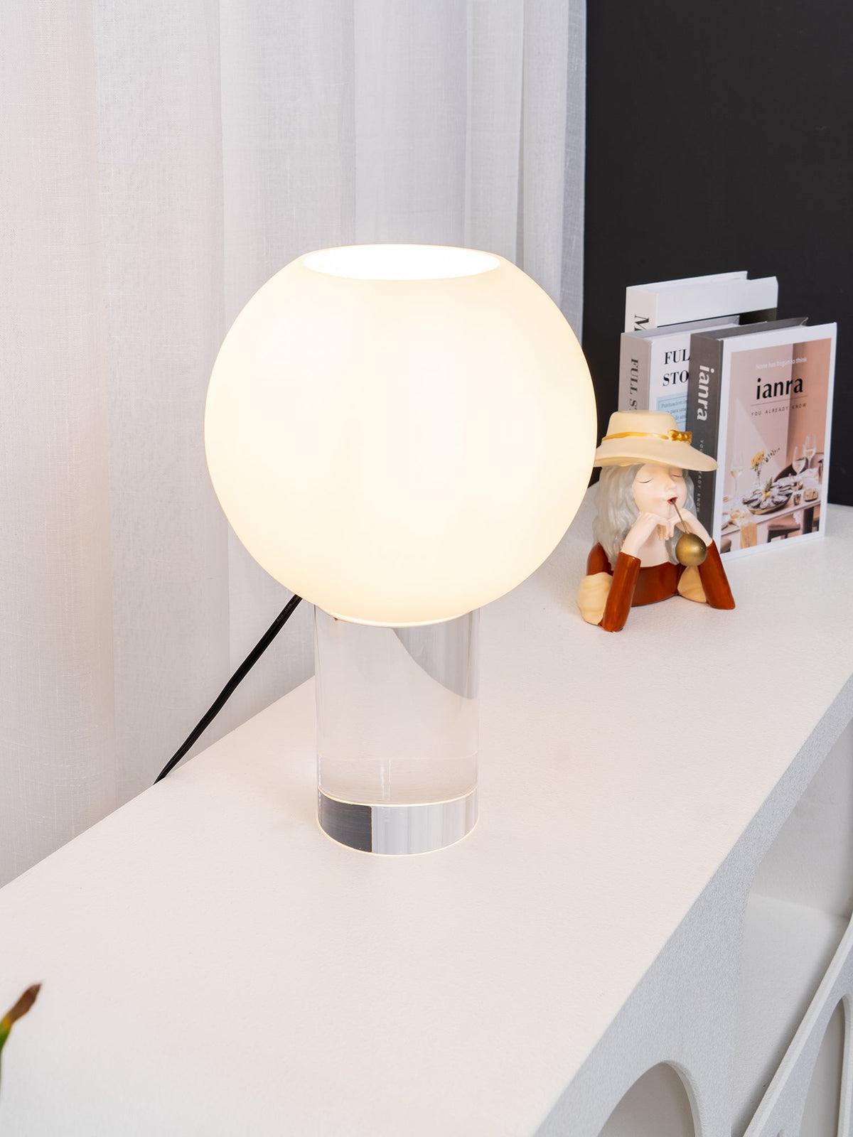 Close on the white glass globe of this Clérachic table lamp, lit, on a white console with a small figurine and books.