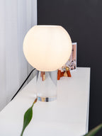 A white glass diffuser lit warm on a clear glass stem, this Clérachic table lamp stands on a white console against a dark wall.