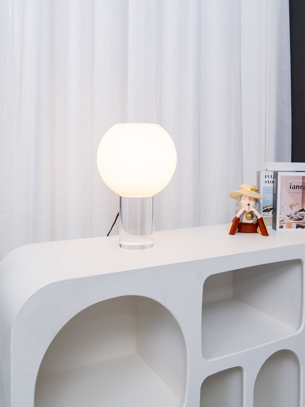 Small Clérachic table lamp, lit, on a white sculpted console with rounded cut-outs, white curtains behind and books to the right.