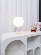 Small Clérachic table lamp, lit, on a white sculpted console with rounded cut-outs, white curtains behind and books to the right.