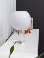 Unlit white glass globe of this Clérachic table lamp on a clear cylindrical base, with a green leaf in the foreground.