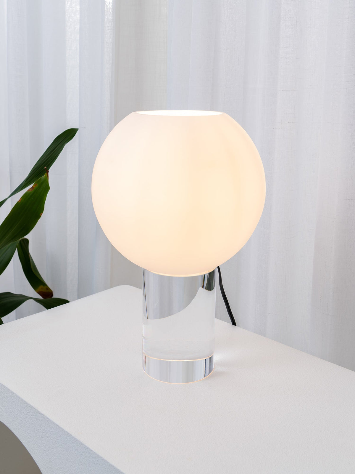 The lit white globe of this Clérachic table lamp rises from a clear glass cylinder, with white curtains and a green leaf close by.