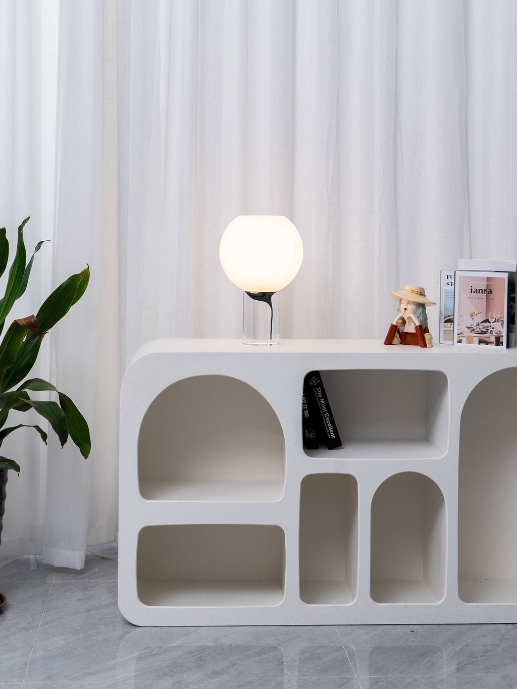 A distant view of a small lit Clérachic table lamp on a white sculpted shelf unit, with curtains and a leafy plant beside it.