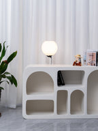 A distant view of a small lit Clérachic table lamp on a white sculpted shelf unit, with curtains and a leafy plant beside it.