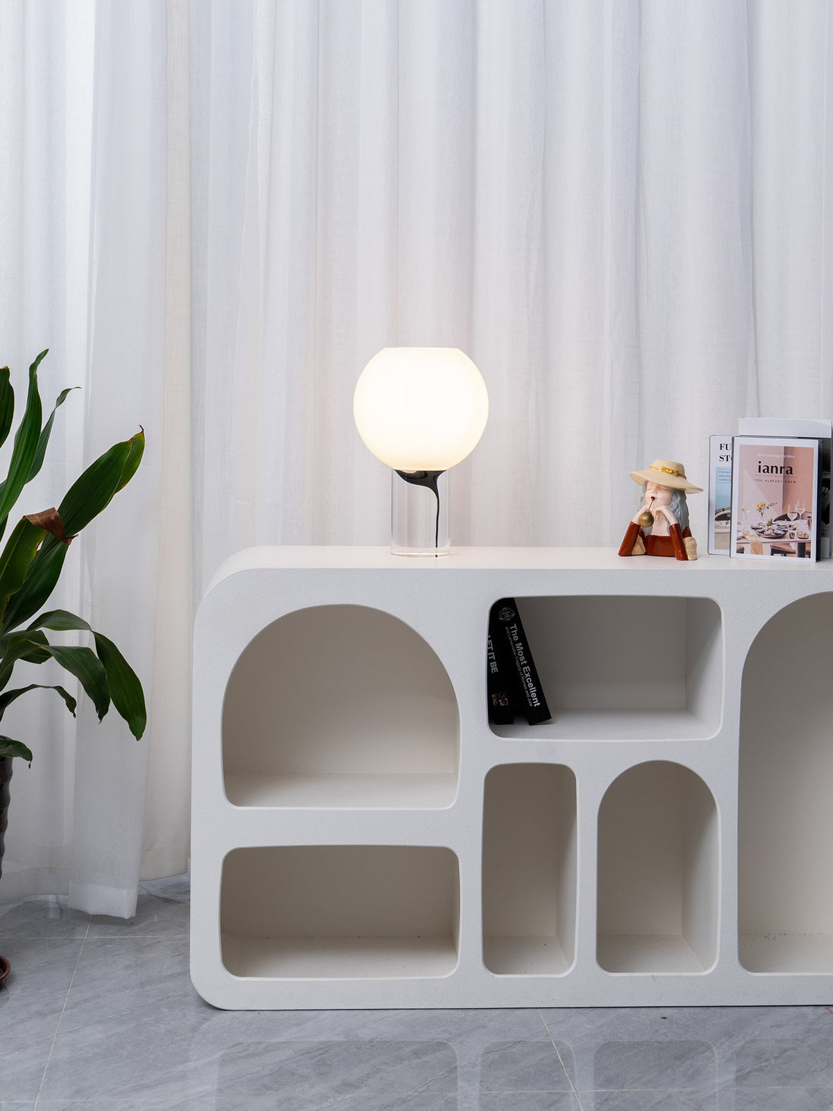 A distant view of a small lit Clérachic table lamp on a white sculpted shelf unit, with curtains and a leafy plant beside it.