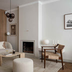 A pale Clérachic globe lamp glows on a white mantel beside two candles, with a woven wooden armchair in a calm, neutral living room.