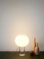 This Clérachic table lamp glows against a pale wall, its glass globe on a clear base, with two tall bottles and a small ball beside it.