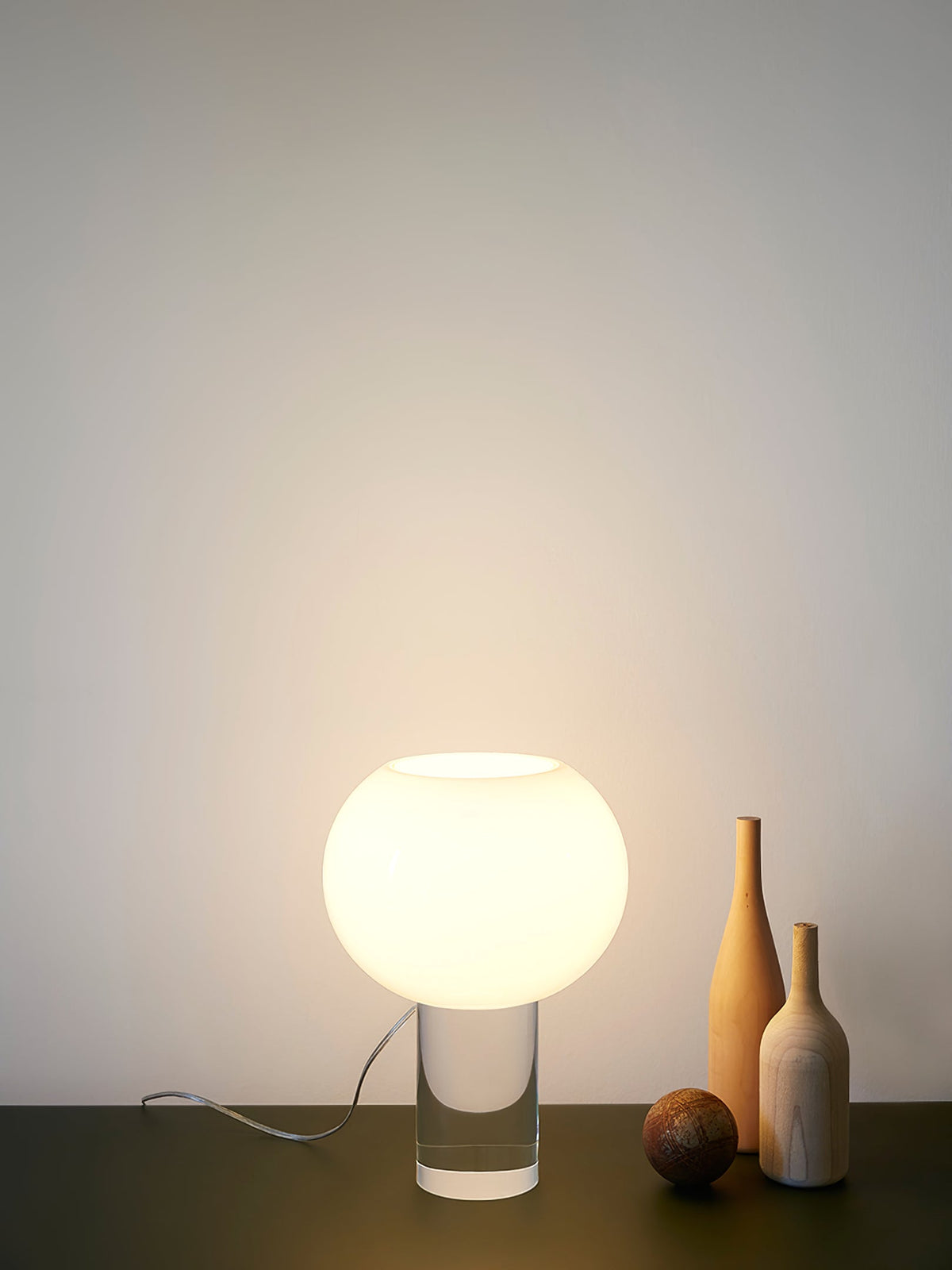 This Clérachic table lamp glows against a pale wall, its glass globe on a clear base, with two tall bottles and a small ball beside it.