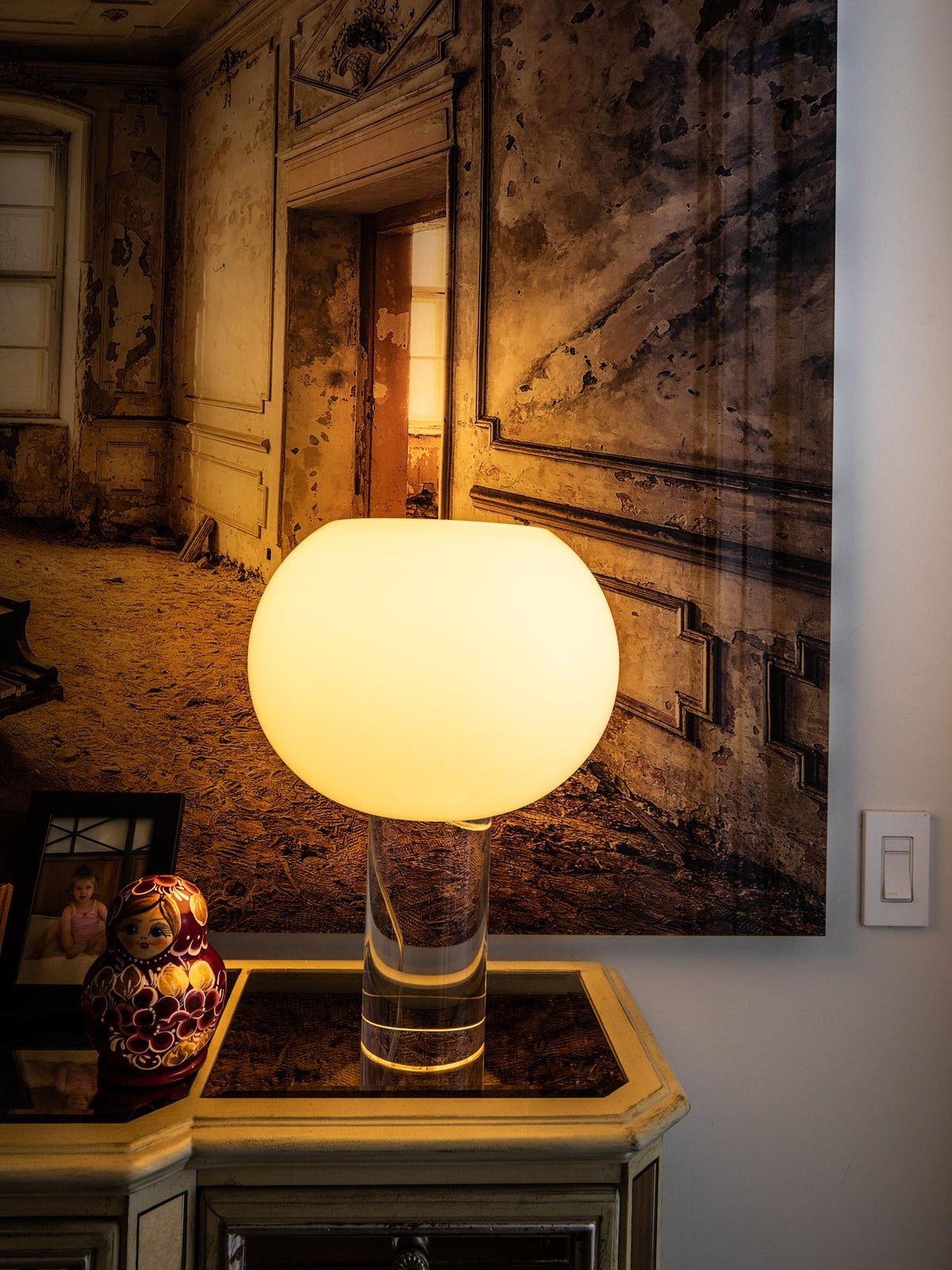 A warm, glowing Clérachic table lamp on a gilt console in front of a large painting of a faded interior, with a nesting doll beside.