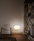 A low, wide Clérachic glass shade glows beside a white armchair in a dim, warm room, with a black metal bookcase at the right.