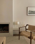 A small Clérachic globe lamp sits on a white ledge beside two candles, near a fireplace and a woven wooden armchair.