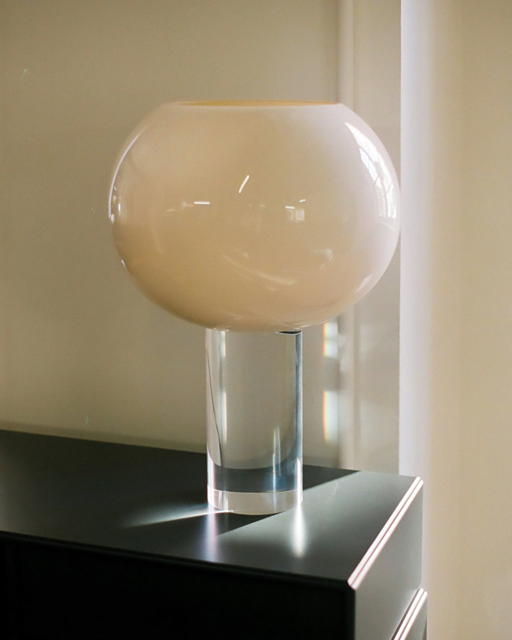 Close view of the cream glass globe on its clear cylindrical stem, this Clérachic table lamp placed on a black cabinet by a window.