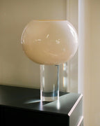 Close view of the cream glass globe on its clear cylindrical stem, this Clérachic table lamp placed on a black cabinet by a window.