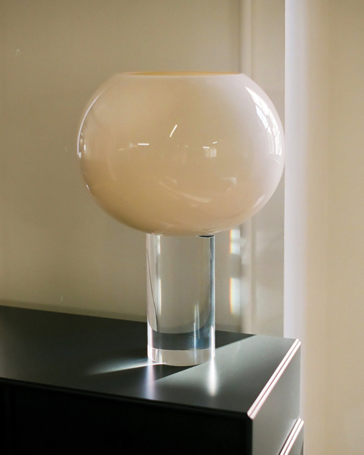 Close view of the cream glass globe on its clear cylindrical stem, this Clérachic table lamp placed on a black cabinet by a window.