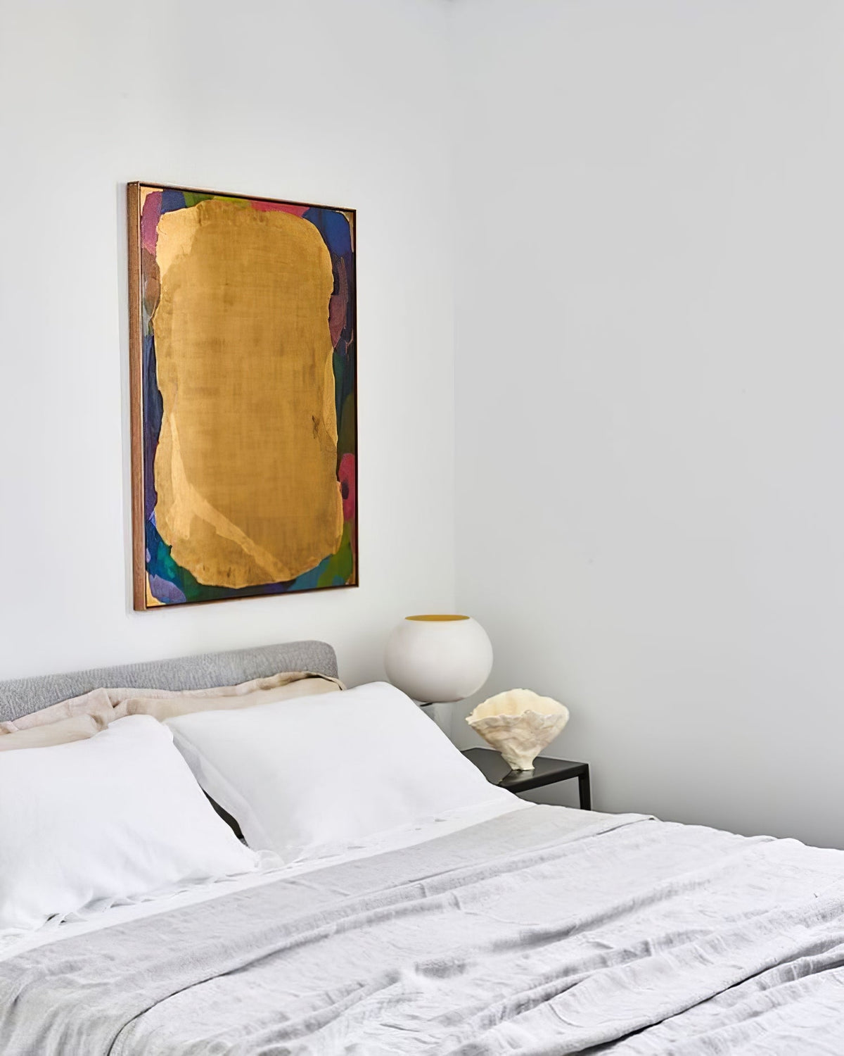 A white Clérachic globe lamp on a bedside table in a calm, pale bedroom, above a bed with white linen and a gold abstract painting.