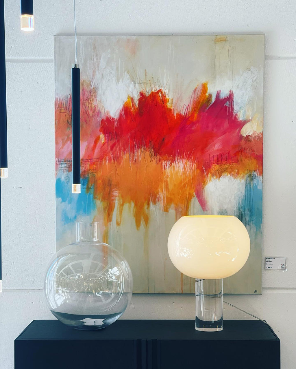 A lit Clérachic table lamp stands on a black shelf beside a large clear glass globe, with a bright abstract painting behind.