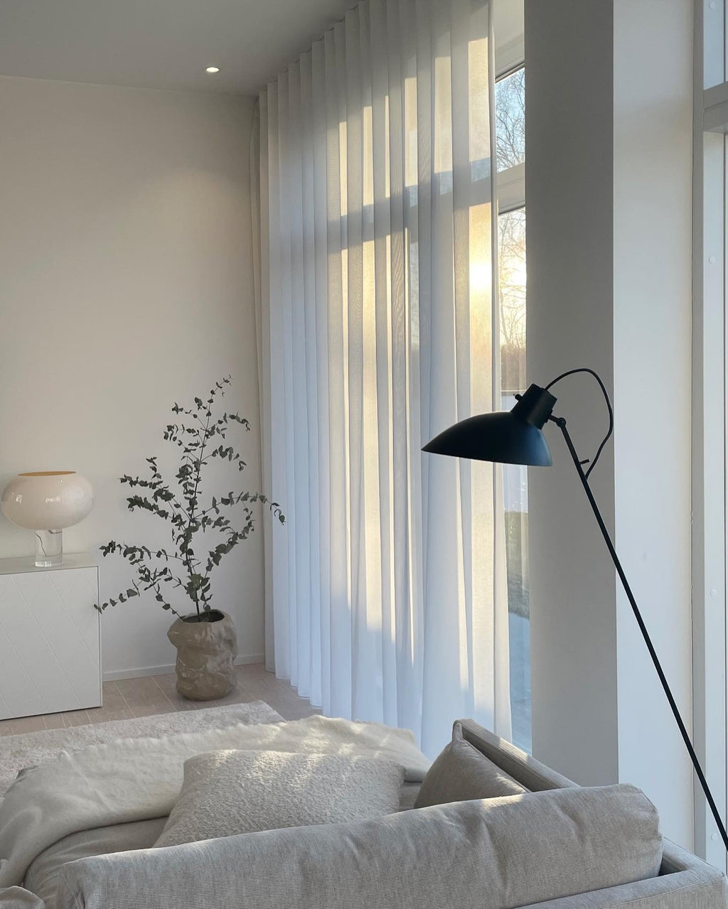 A soft-lit living room with a white glass Clérachic lamp on a white cabinet, a potted eucalyptus, a black floor lamp and a pale sofa.