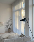 A soft-lit living room with a white glass Clérachic lamp on a white cabinet, a potted eucalyptus, a black floor lamp and a pale sofa.