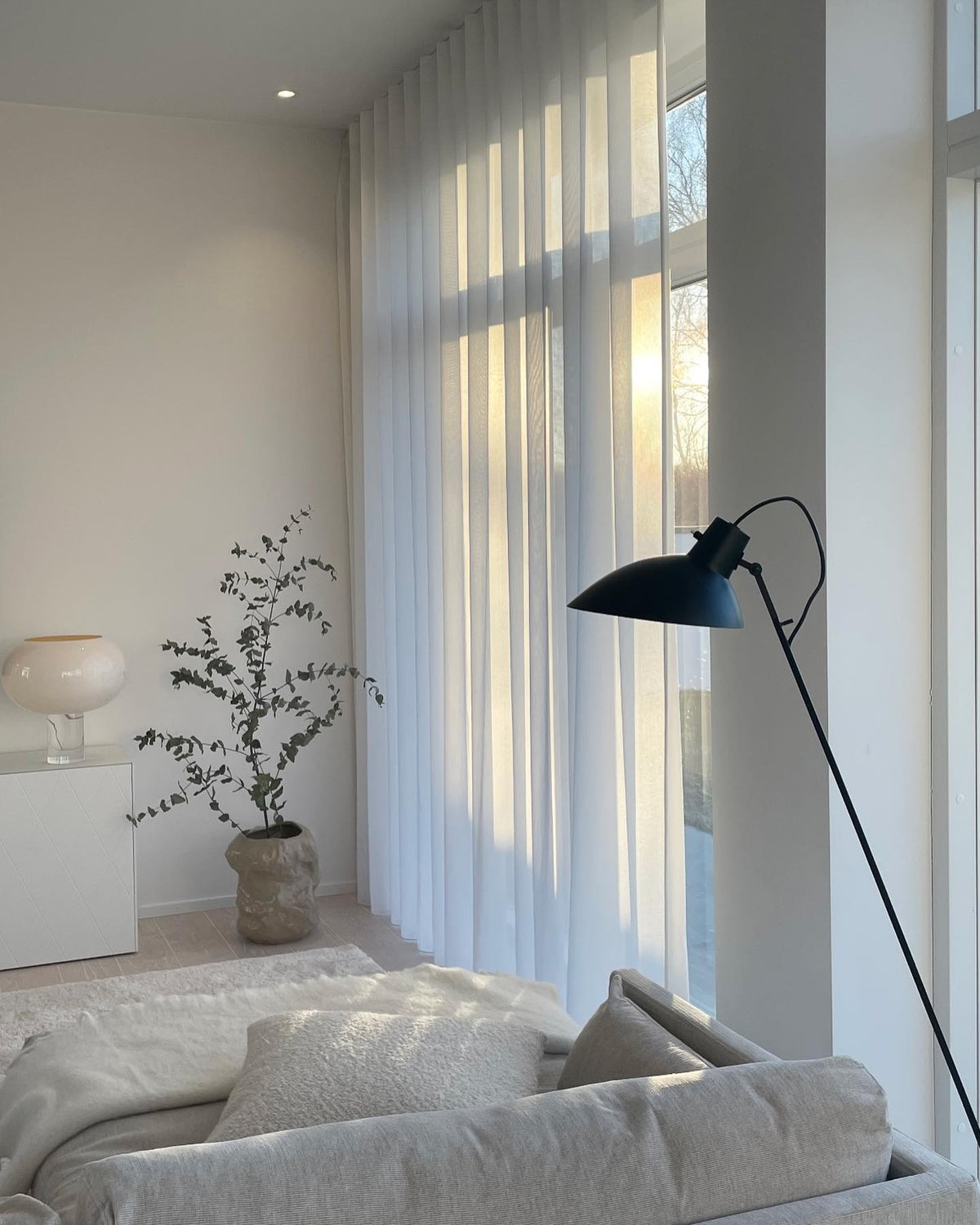 A soft-lit living room with a white glass Clérachic lamp on a white cabinet, a potted eucalyptus, a black floor lamp and a pale sofa.
