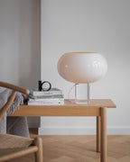 A Clérachic table lamp with a wide white glass shade on a clear base stands on a pale wooden desk beside a stack of books.