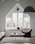 A small Clérachic glass table lamp sits on the window ledge of a bright attic room with a round coffee table and a dark pendant.