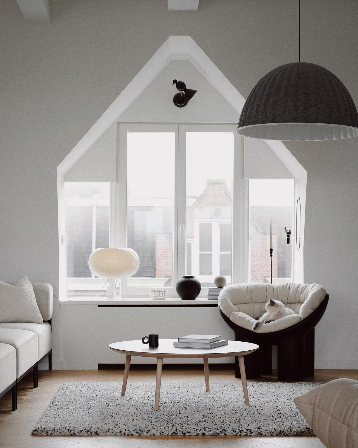 A small Clérachic glass table lamp sits on the window ledge of a bright attic room with a round coffee table and a dark pendant.