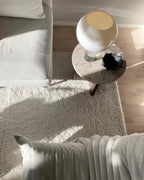 Seen from above, a white glass Clérachic globe lamp stands on a round marble side table beside a cream sofa and a shaggy rug.