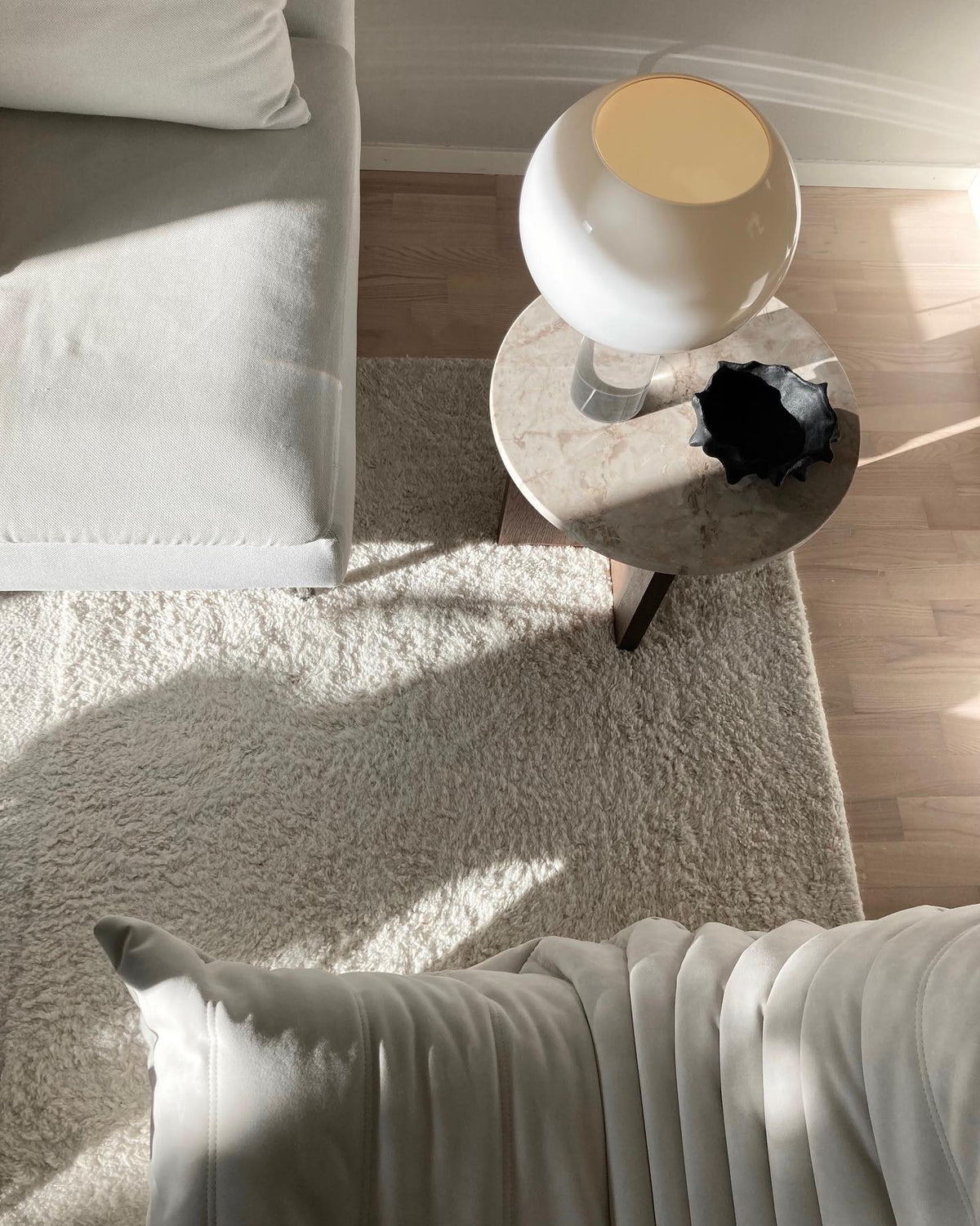Seen from above, a white glass Clérachic globe lamp stands on a round marble side table beside a cream sofa and a shaggy rug.
