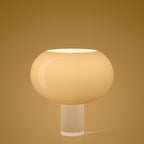 A glowing Clérachic table lamp with a wide, rounded glass shade on a clear cylinder, on a warm gold backdrop.