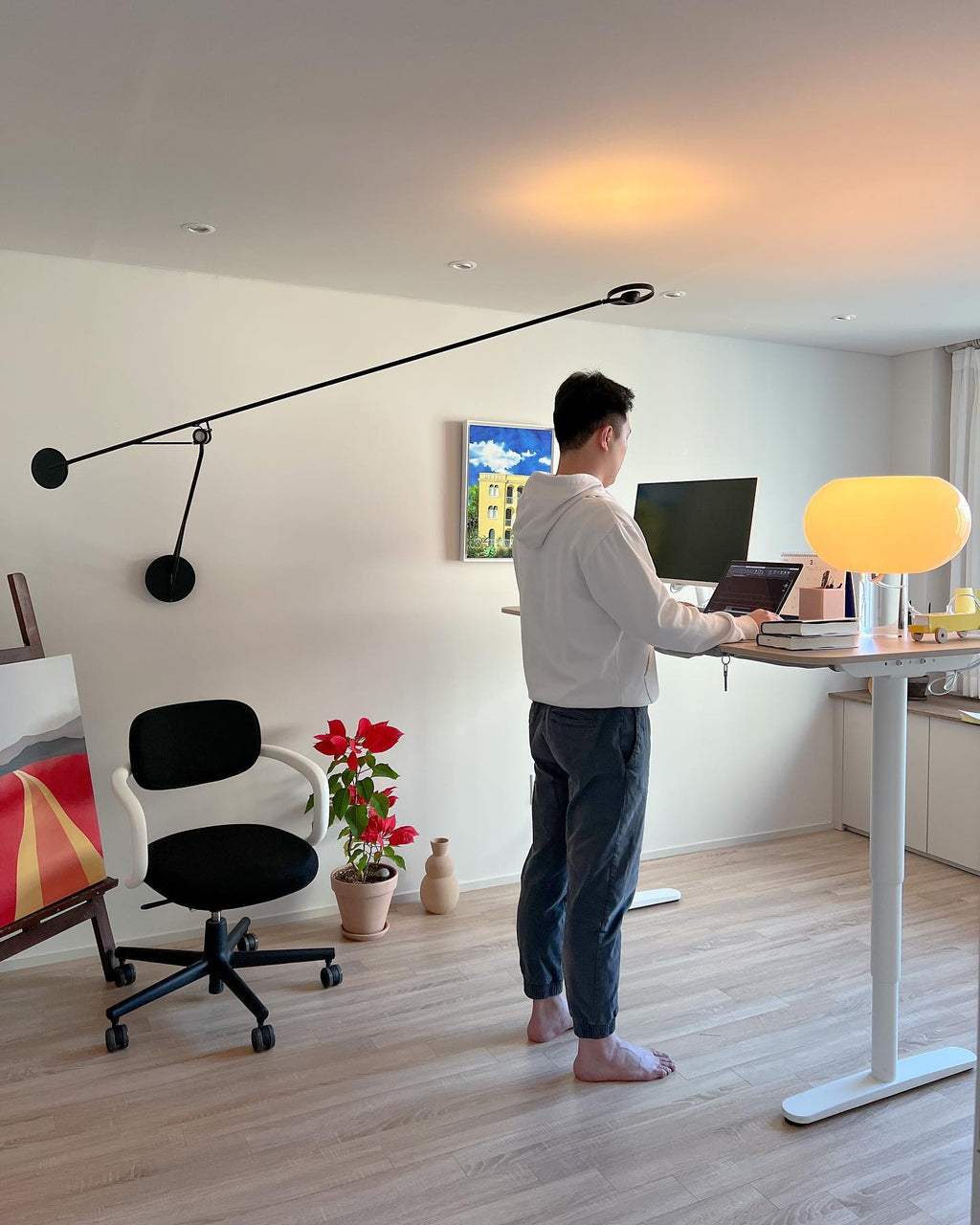 A lit Clérachic glass table lamp glows on a pale wooden standing desk, with a man at the laptop and a black office chair behind.