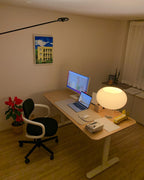 A lit Clérachic table lamp on a light wooden desk beside a laptop and monitor, with a red plant and a black chair nearby.