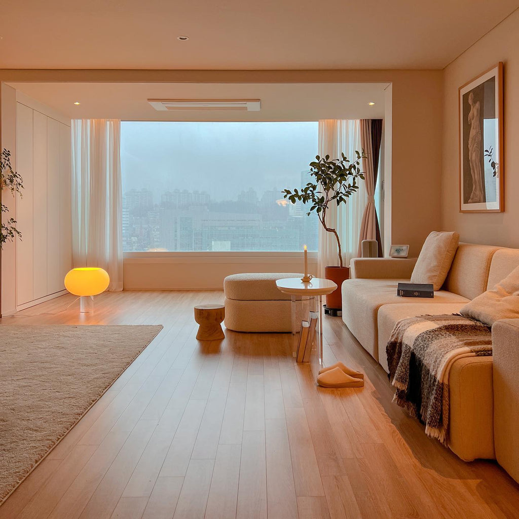 A small amber-lit Clérachic lamp glows on the wooden floor of a wide living room, with a big window, a sofa and a throw beyond.
