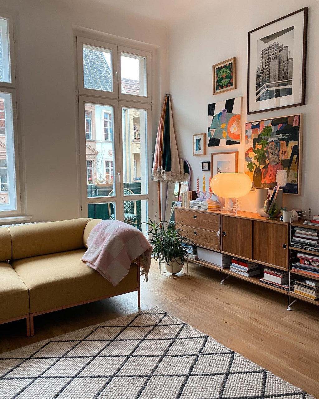 A lit Clérachic table lamp on a walnut sideboard in a bright room with framed prints, a yellow sofa and tall windows.
