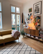 A lit Clérachic table lamp on a walnut sideboard in a bright room with framed prints, a yellow sofa and tall windows.