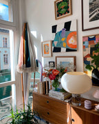 Close on a lit white glass Clérachic table lamp on a walnut sideboard, with a vase of red flowers and a wall of colourful art above.