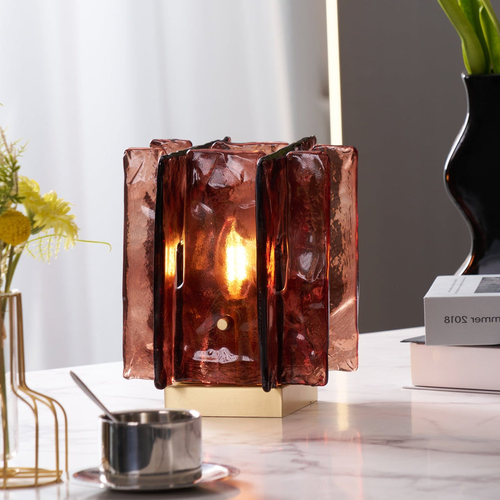 A lit Clérachic table lamp with textured amber-red glass panels glows on a marble tabletop, beside a steel cup and a vase of yellow flowers.